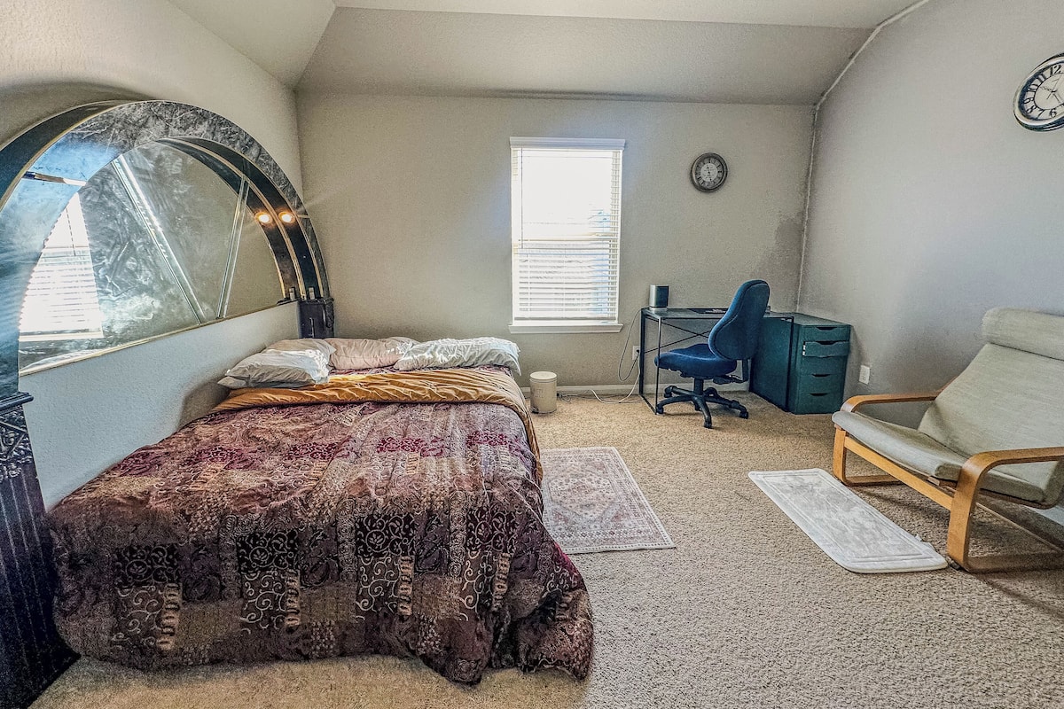 A bedroom with a bed, a desk, a chair, a mirror, a clock, and a window.