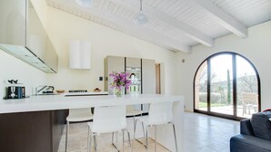 A modern kitchen with a white countertop, bar stools, and a view of the outdoors through a large window. - Luxurious home in Trequanda with outdoor swimming pool (Trequanda)