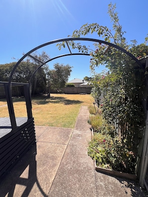 A backyard with a wooden fence, a concrete pathway, and a metal archway. - Warrnambool Holiday Home (Warrnambool)