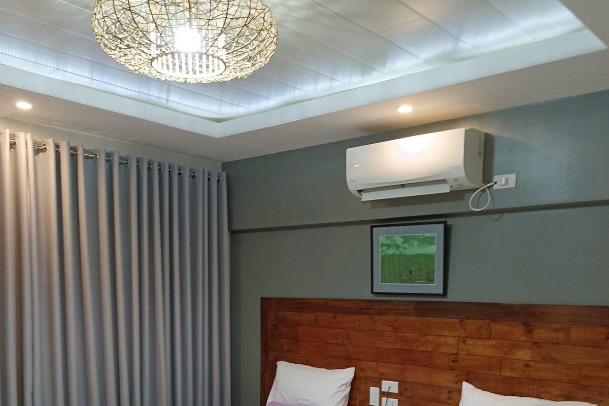 Ein Hotelzimmer mit zwei Einzelbetten, Holz-Kopfteilen, einer Deckenleuchte, einer Klimaanlage und einem gerahmten Bild an der Wand.