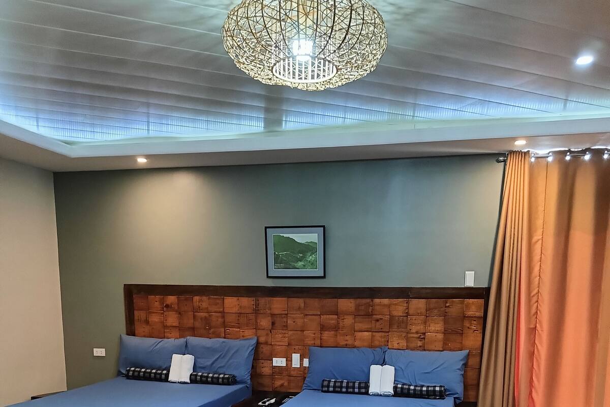 Ein Zimmer mit zwei Holzbetten, einem hölzernen Kopfteil, einer Deckenleuchte und einem gerahmten Bild an der Wand.