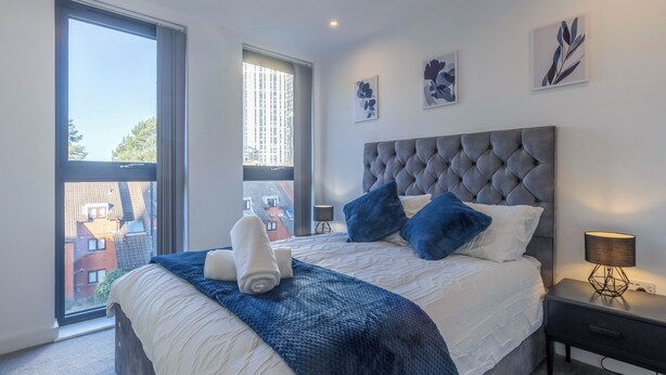 A modern bedroom with a large bed, a grey tufted headboard, and a view of buildings through a large window. - A classy, cozy & modern apartment in Birmingham City centre. (Birmingham)