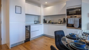 A modern kitchen with white cabinetry, a built-in oven, and a dining area with a glass table and blue bowls. - A classy, cozy & modern apartment in Birmingham City centre. (Birmingham)
