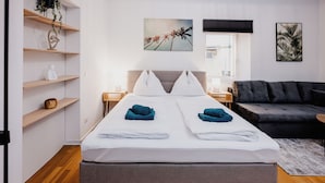 A modern hotel room with a double bed, two nightstands, a grey sofa, wooden shelves, and artwork on the walls.