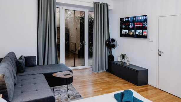 A modern living room with a grey sectional sofa, a small round table, a black side table with a fan, and a TV mounted on the wall displaying movie posters.