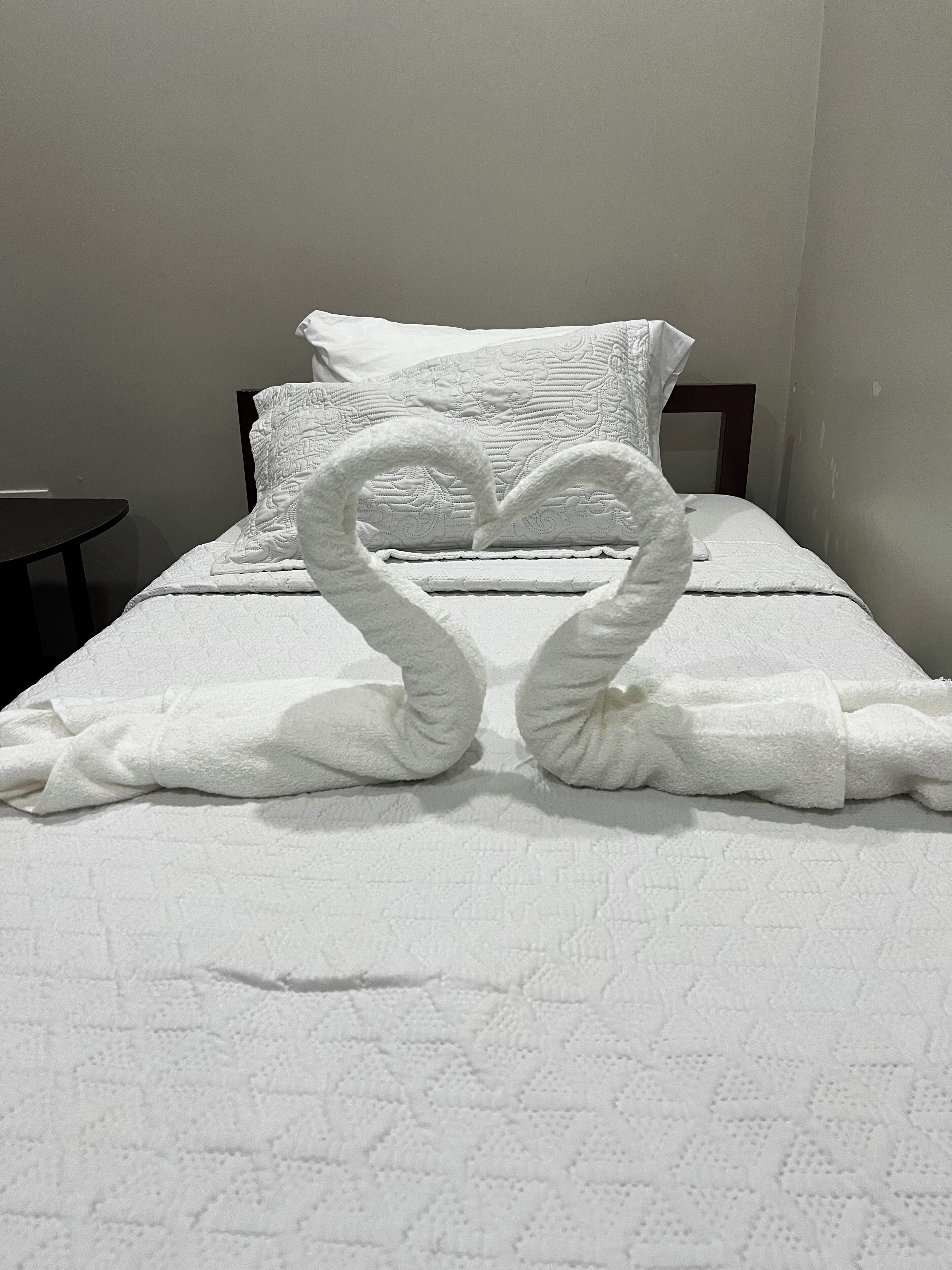 A bed with a white quilt and pillows, and two towels arranged in a heart shape on the bed.