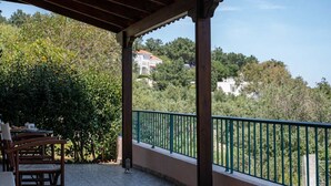 Uma área externa coberta com uma mesa e cadeiras, um corrimão verde e vista para árvores e edifícios ao fundo.