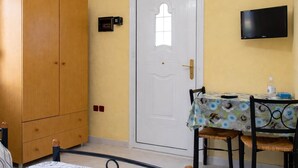 Quarto com guarda-roupa de madeira, porta branca com janela, mesa com toalha de mesa florida e televisão na parede.
