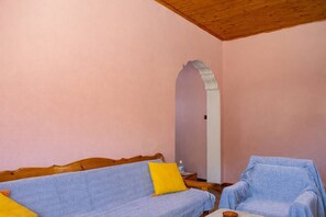 A room with a wooden ceiling, a pink wall, a sofa with a patterned cover, a chair with a blue cover, and a small table with a bottle.