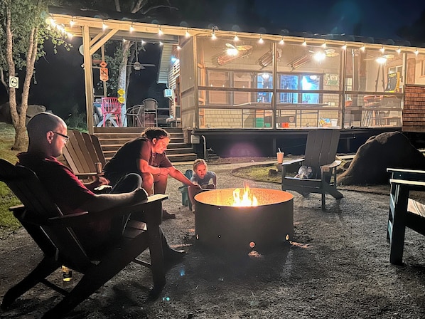 A nighttime outdoor seating area with a fire pit, chairs, and a covered structure with string lights.