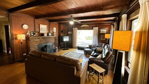 A cozy living room with a fireplace, leather sofas, a television, and a large window with curtains. - Deer Hollow Cabin - A family-friendly 3-bedroom cabin in charming Three Rivers (Three Rivers)
