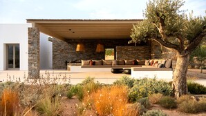Una casa moderna con fachada de piedra, un área de estar exterior techada con un sofá y cojines, y un árbol en primer plano.