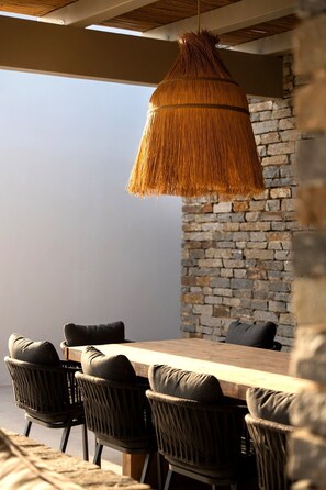 Un coin repas avec une table en bois et des chaises en osier, un luminaire suspendu en rotin et un mur en pierre en arrière-plan.