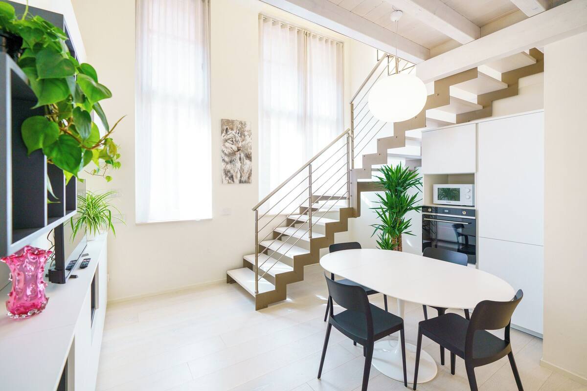 A modern kitchen with a white table and chairs, a staircase, and a potted plant.