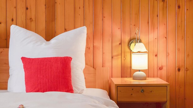 Ein Bett mit weißen und roten Kissen, ein Nachttisch aus Holz mit einer Lampe und eine Holzvertäfelung an der Wand.