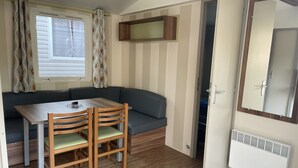 Una habitación pequeña con una mesa y sillas de madera, un sofá gris, una ventana con cortinas, un espejo y un radiador.