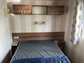 A bedroom with a bed, a wooden headboard, a wall-mounted shelf, a bedside table, and a window with curtains. - Mobile home in the heart of the Homair la pointe du Médoc campsite (Le Verdon-sur-Mer)