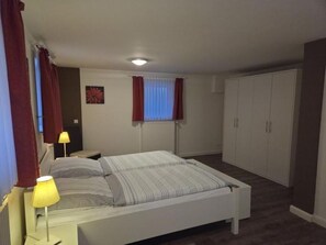 Ein Schlafzimmer mit Bett, Nachttischlampen, Heizkörper, einem Fenster mit Vorhängen und einem Kleiderschrank.