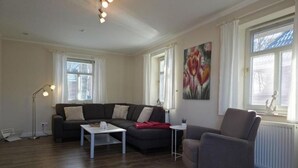 A living room with a dark-colored sofa, a white coffee table, and a grey armchair. There is a painting on the wall and a window with blinds. - Plenty of space for the whole family - North Sea vacation at Haus Küstenwind - House (Wyk auf Föhr)