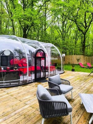Eine transparente Kuppelkonstruktion mit rotem Innenraum, umgeben von einer Holzterrasse und Gartenmöbeln.