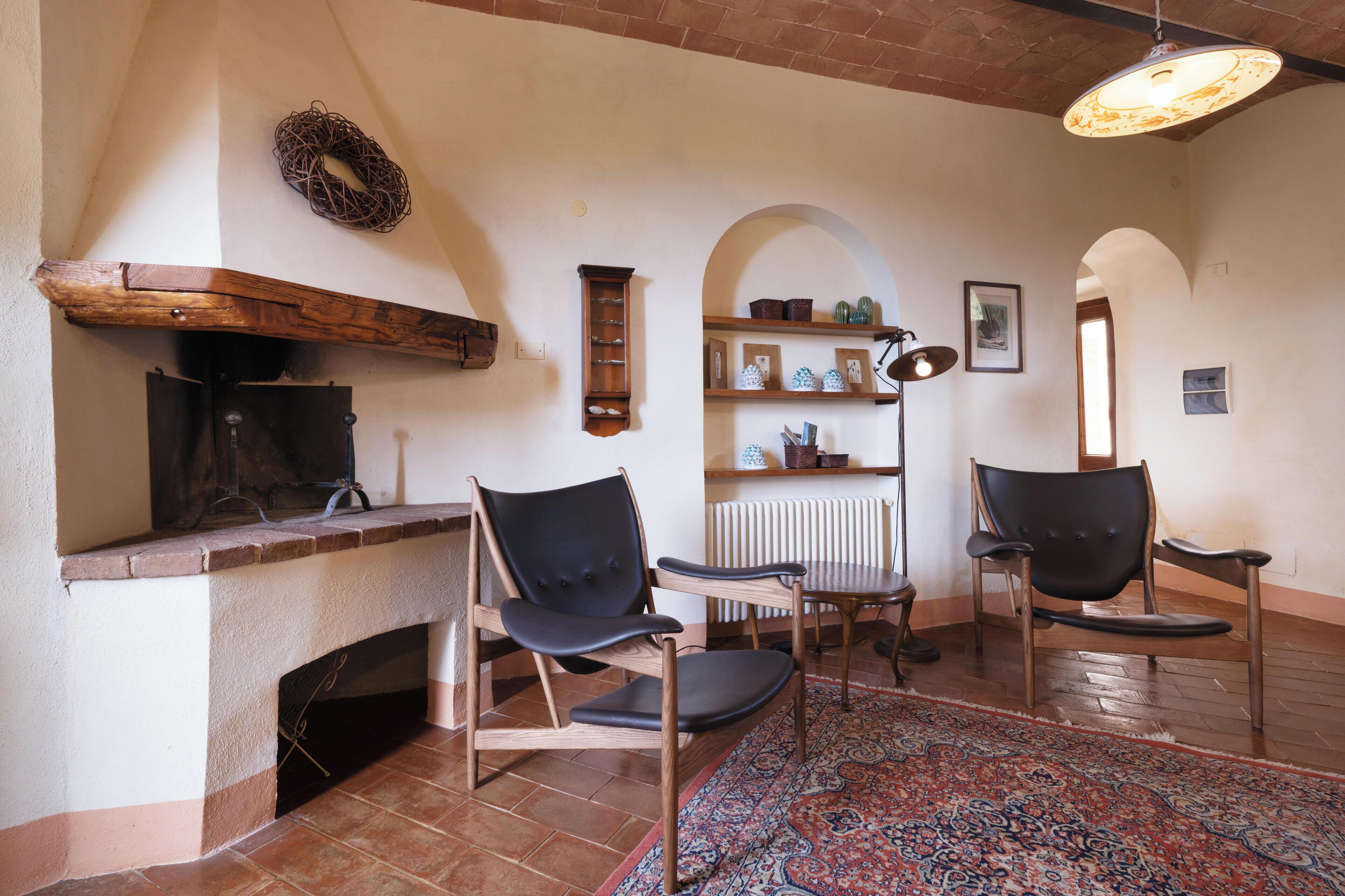 A cozy living room with a fireplace, two chairs, a patterned rug, and a shelf with decorative items.