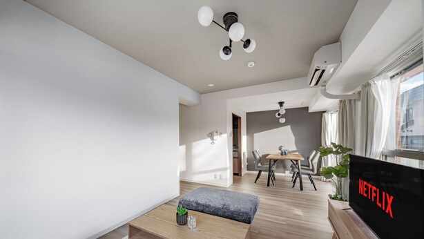 A modern living room with a wooden coffee table, a grey sofa, a TV displaying the Netflix logo, and a dining area in the background. - E-US NAEBO 203 (Sapporo)