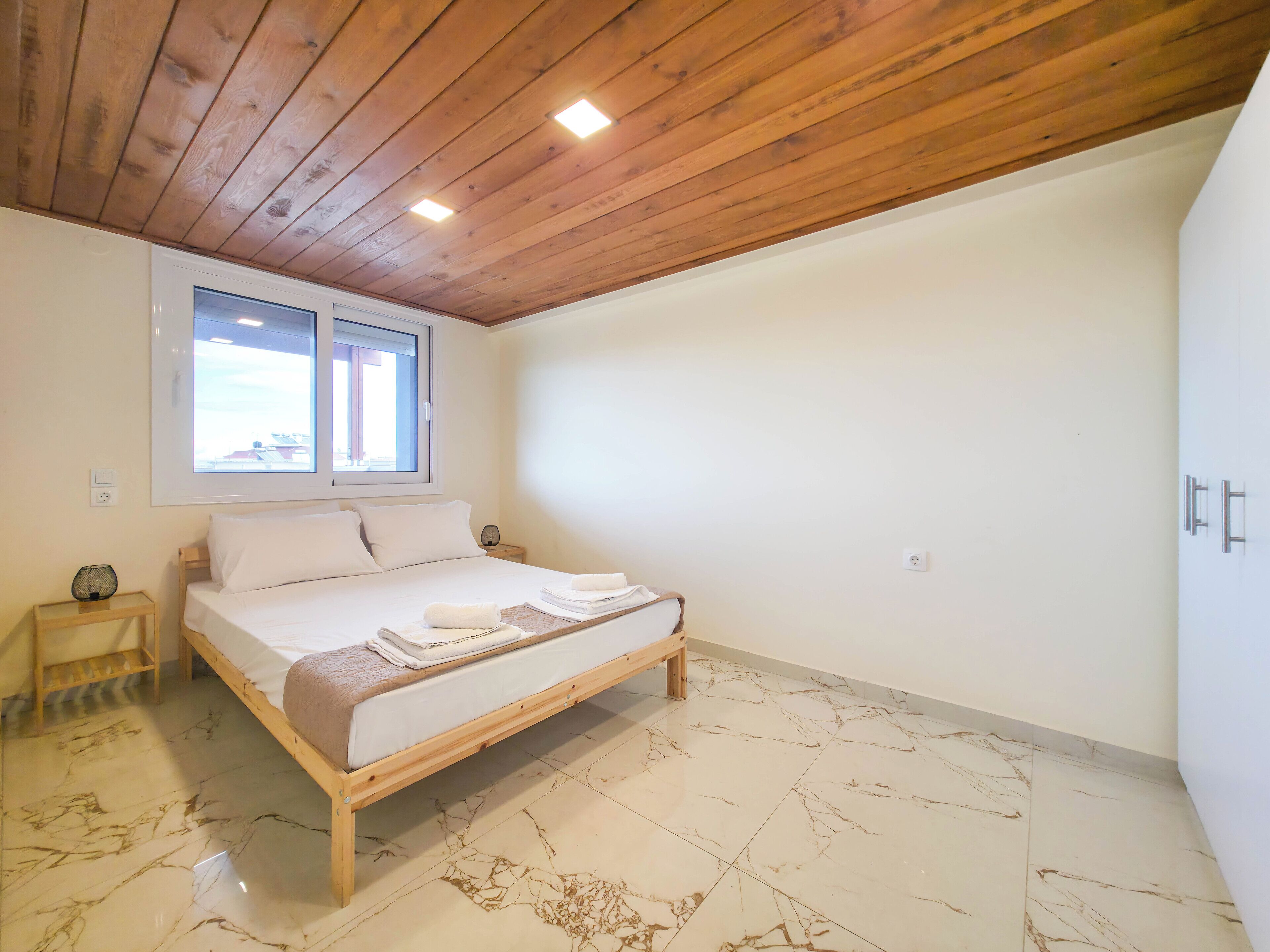 A bedroom with a wooden ceiling, a bed with white linens, a small bedside table, and a window with a view.