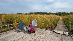 A wooden boardwalk leading through a field of tall grass, with a blue chair and a white chair with a pink blanket on it. - Seaside plot with private jetty, rowing boat & sunset (Kvicksund)