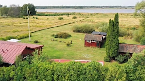 A rural property with a large open field, a small wooden house, and a tall coniferous tree. - Seaside plot with private jetty, rowing boat & sunset (Kvicksund)
