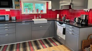 A modern kitchen with grey cabinets, a black countertop, and a red backsplash. There is a window with a view of greenery outside, a sink, a microwave, and a coffee maker. - Seaside plot with private jetty, rowing boat & sunset (Kvicksund)