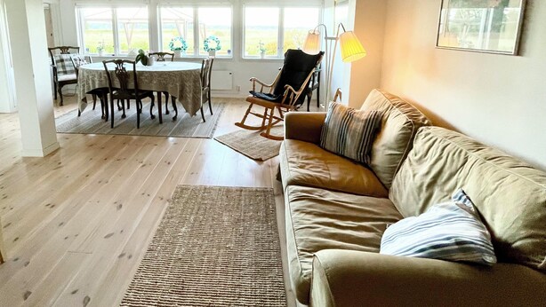 A spacious living room with a wooden ceiling, a beige sofa, a dining table with chairs, and a view through large windows. - Seaside plot with private jetty, rowing boat & sunset (Kvicksund)
