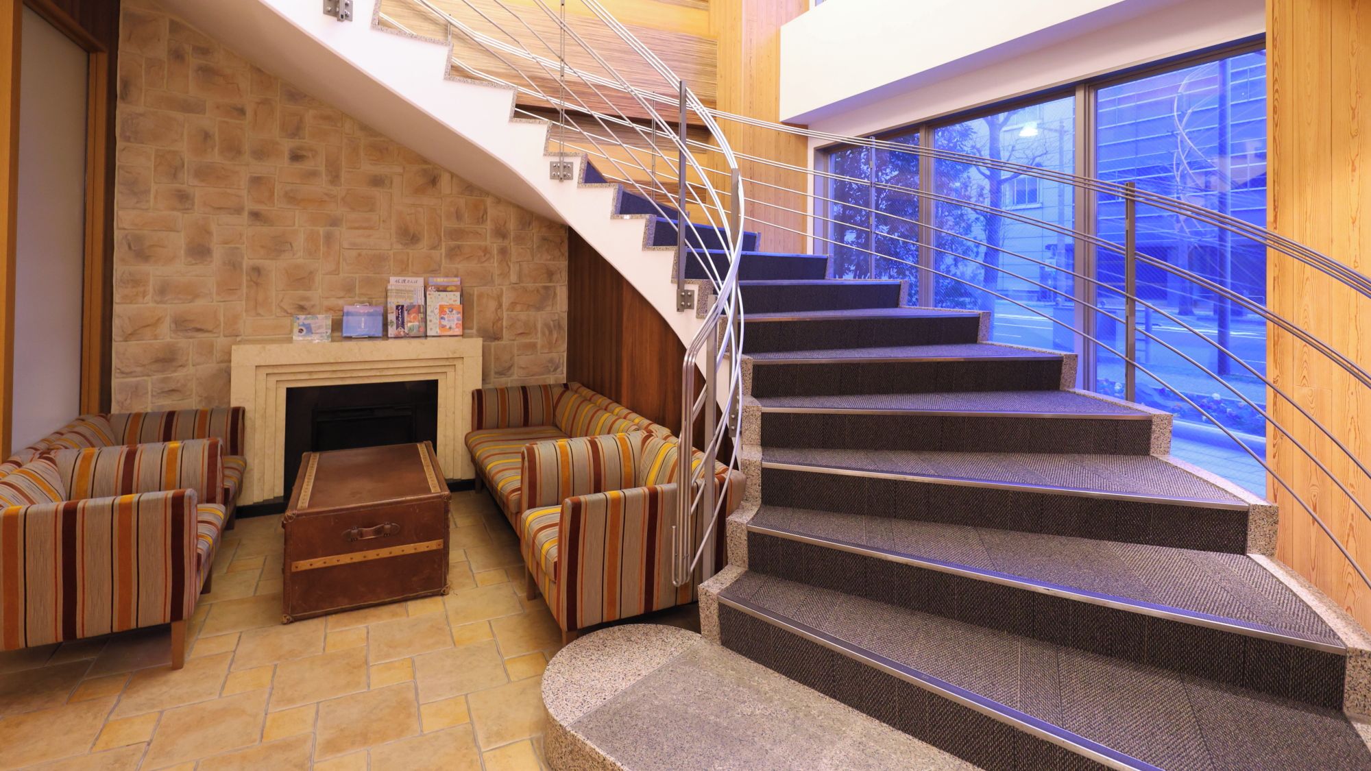 A modern interior with a stone fireplace, striped sofas, a wooden chest, and a spiral staircase.