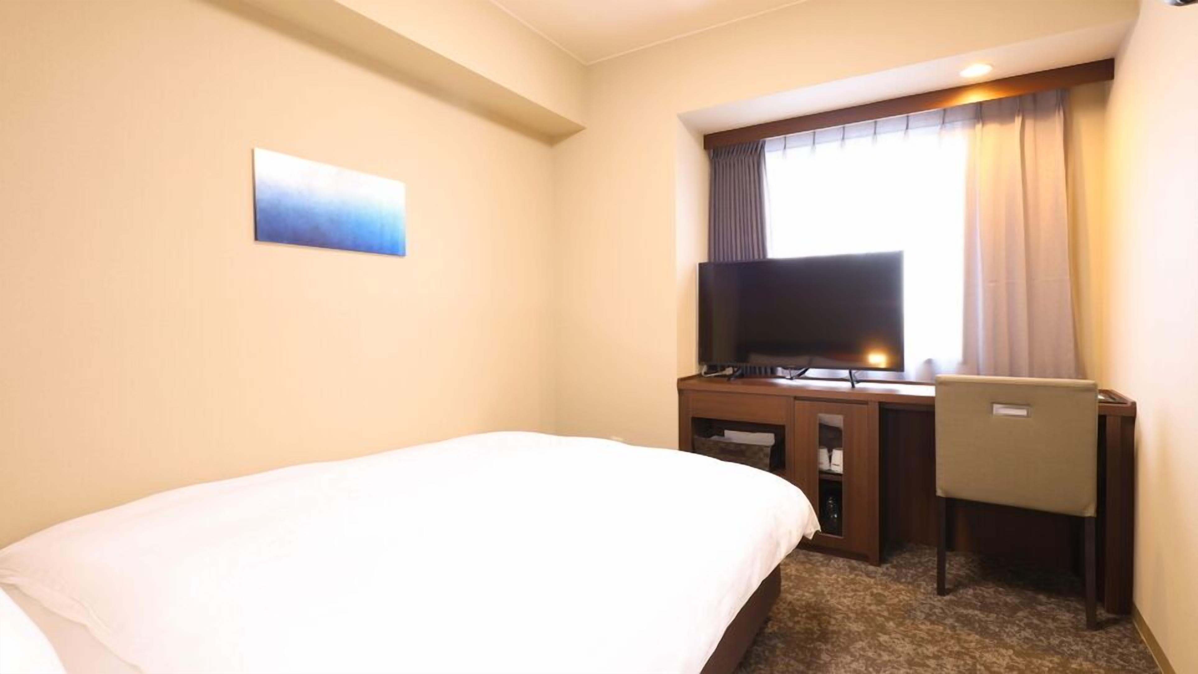 A hotel room with a bed, a desk with a television, and a window with curtains.