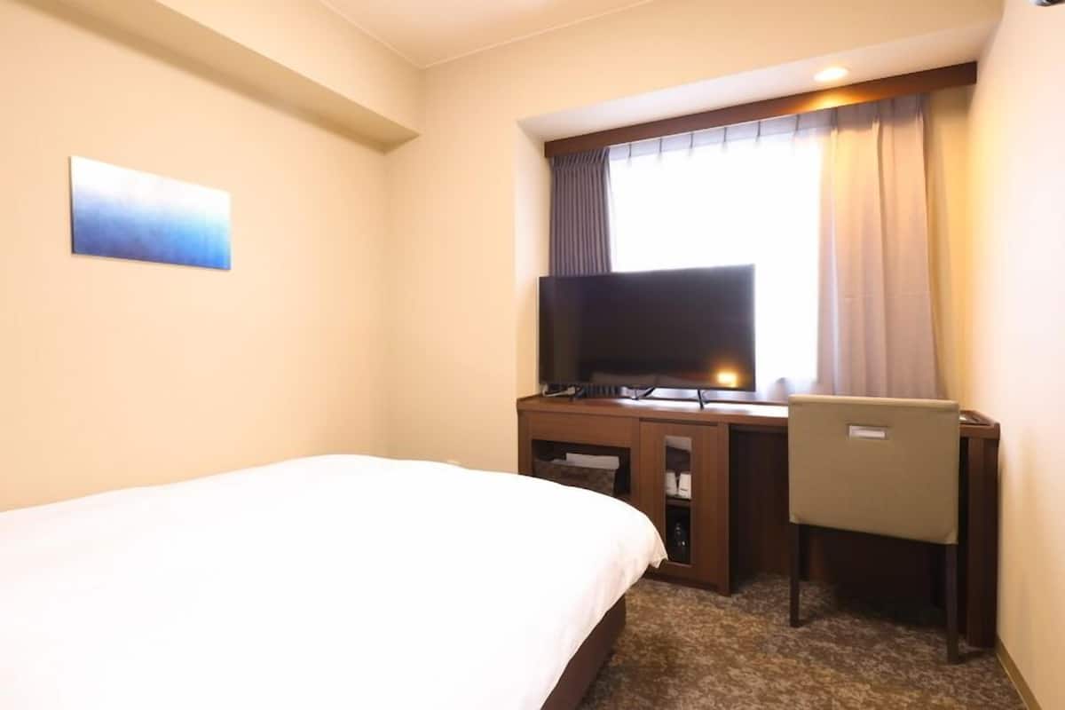 A hotel room with a bed, a desk with a television, and a window with curtains.