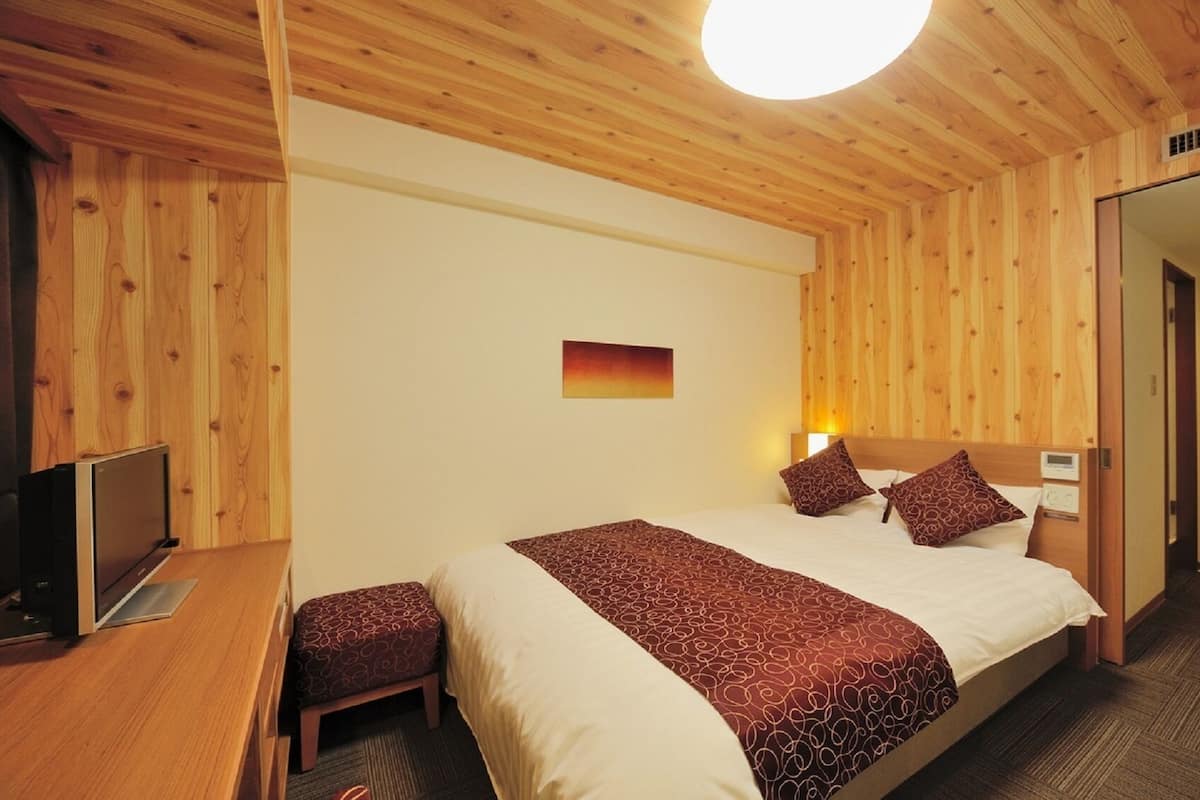 A hotel room with a wooden ceiling, a bed with patterned bedding, a bedside table, a TV, and a small stool.