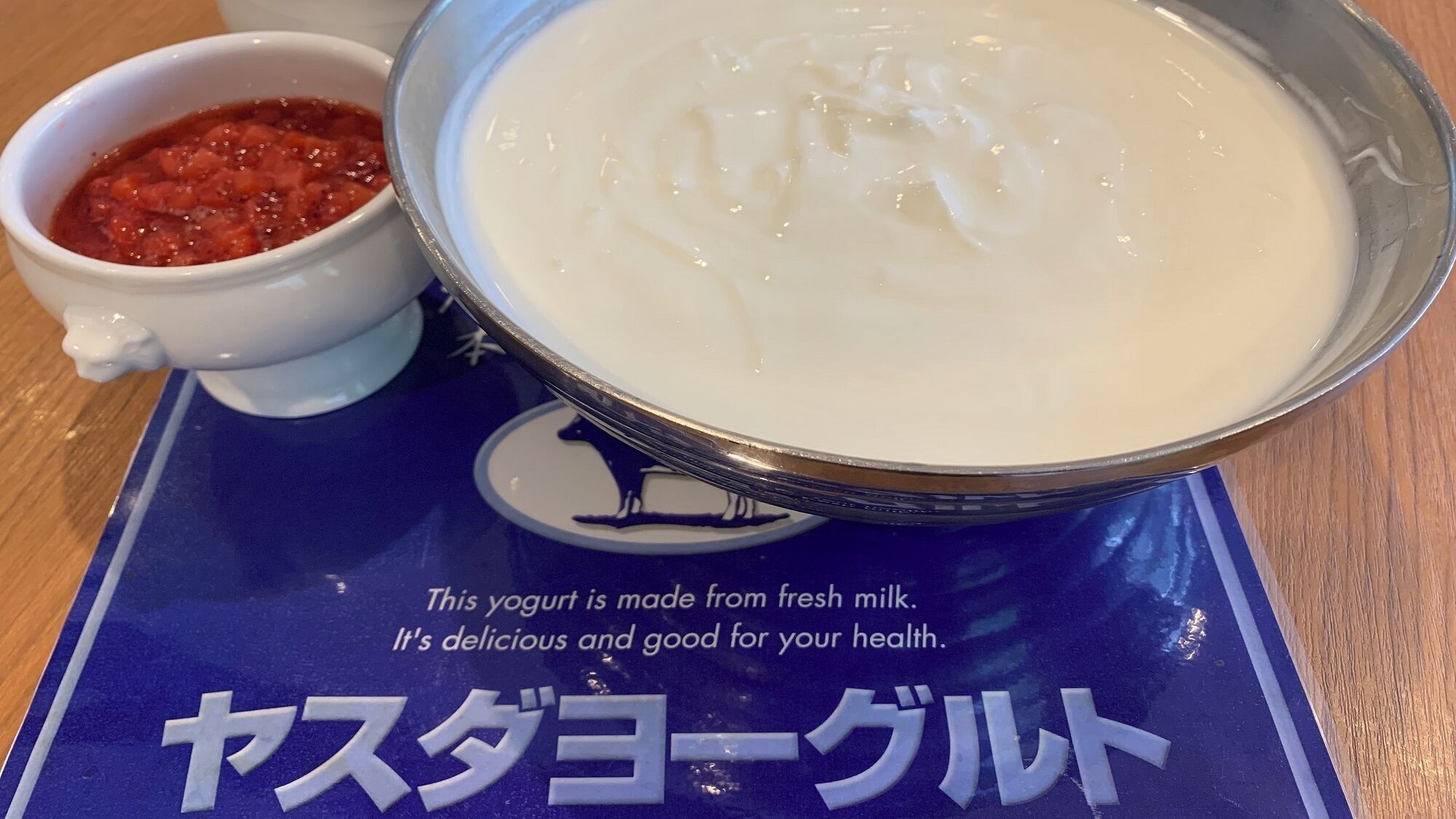 Un tazón de yogur con salsa roja al lado, sobre un mantel individual azul con texto en japonés.