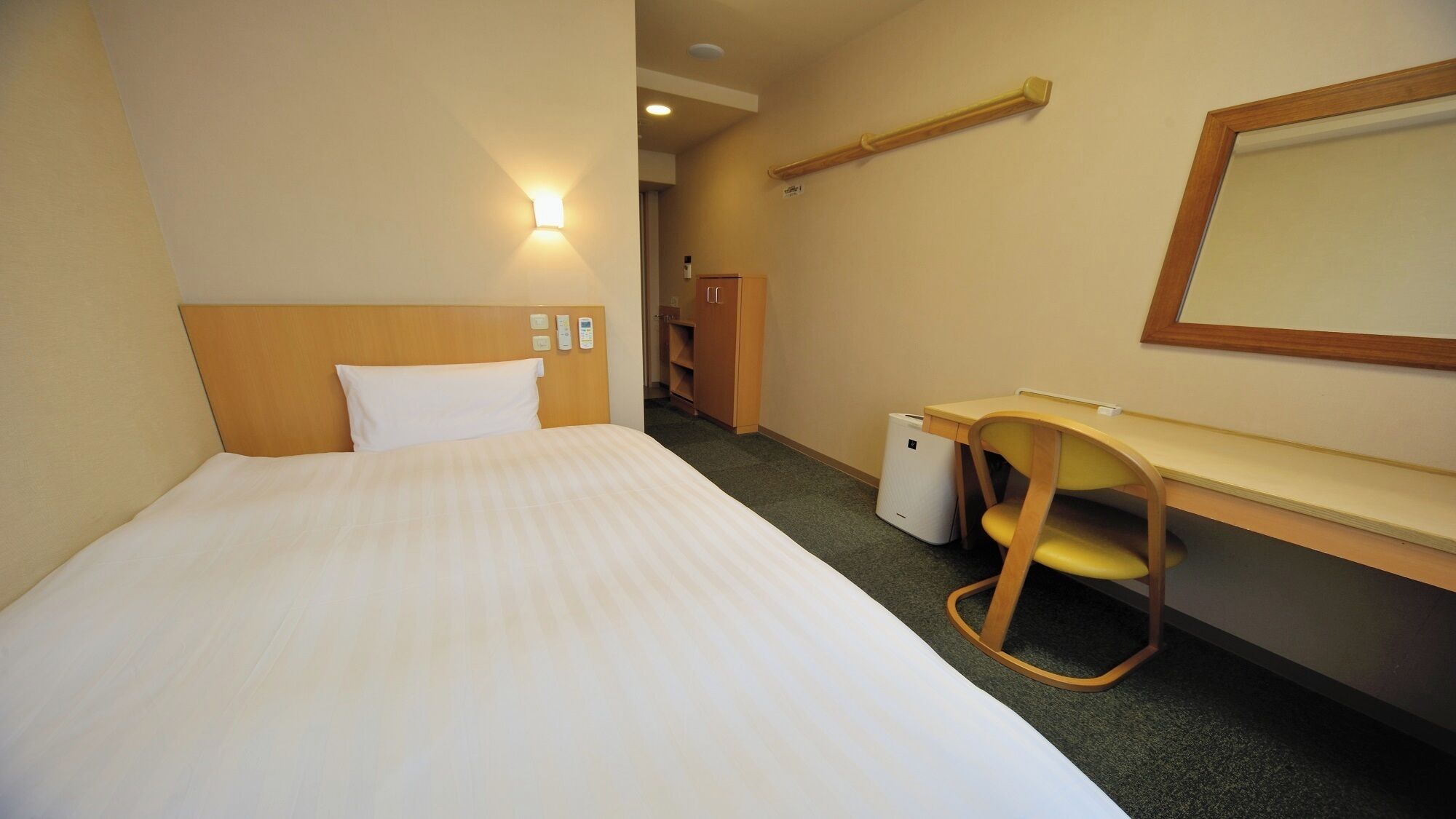 A hotel room with a bed, a desk, a chair, a small trash bin, and a mirror.