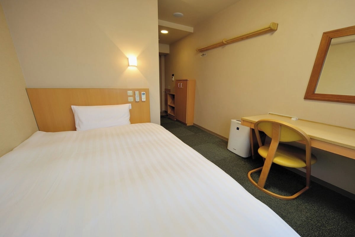 A hotel room with a bed, a desk, a chair, a small trash bin, and a mirror.