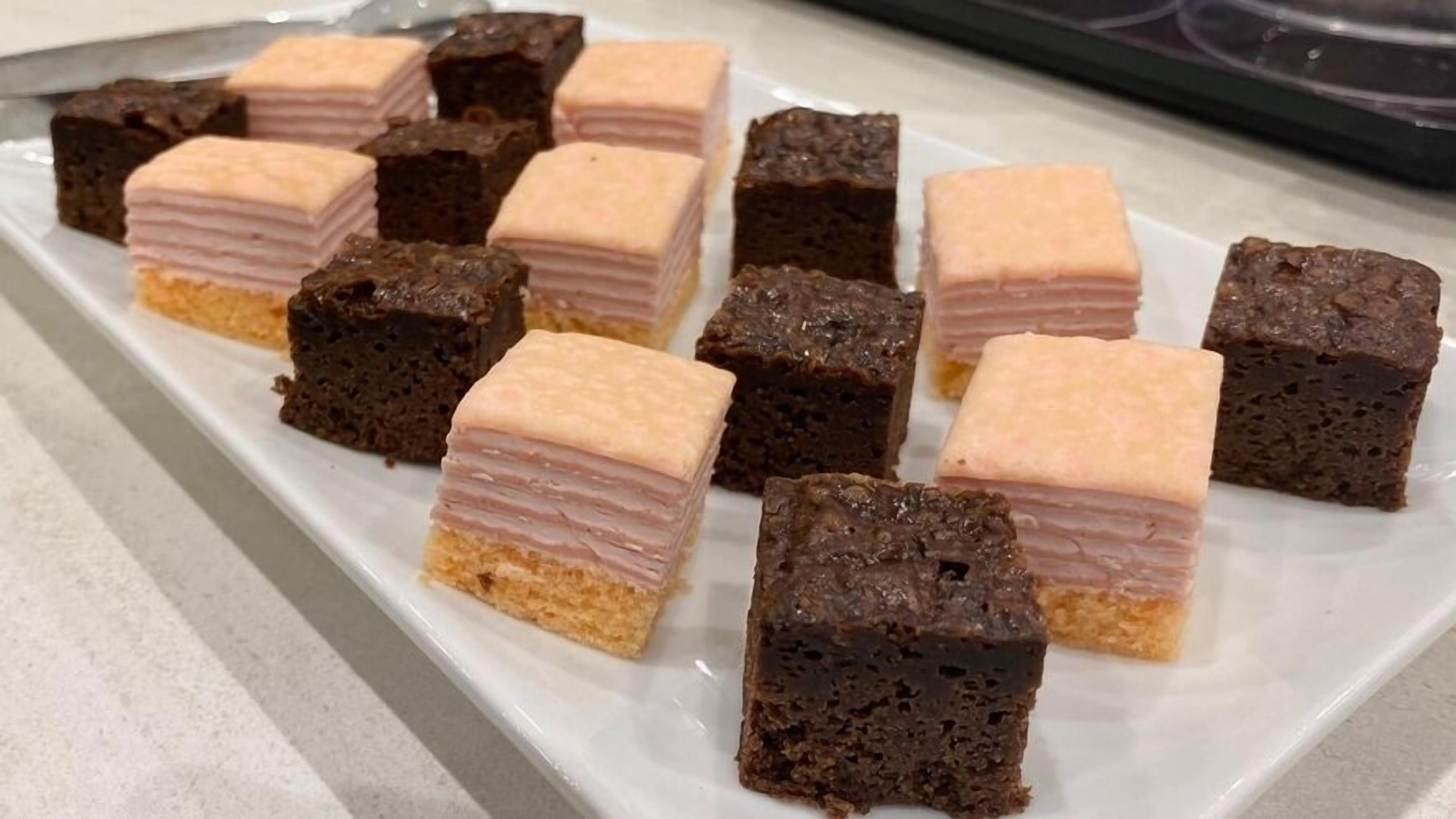 A rectangular plate with several square-shaped desserts, some with pink layers on top.