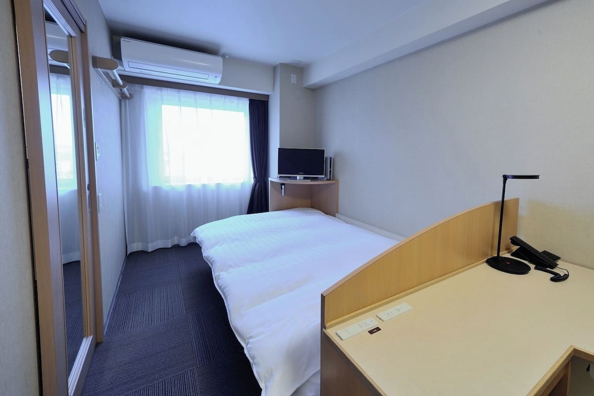 A hotel room with a bed, a desk with a telephone, a television, and an air conditioning unit.