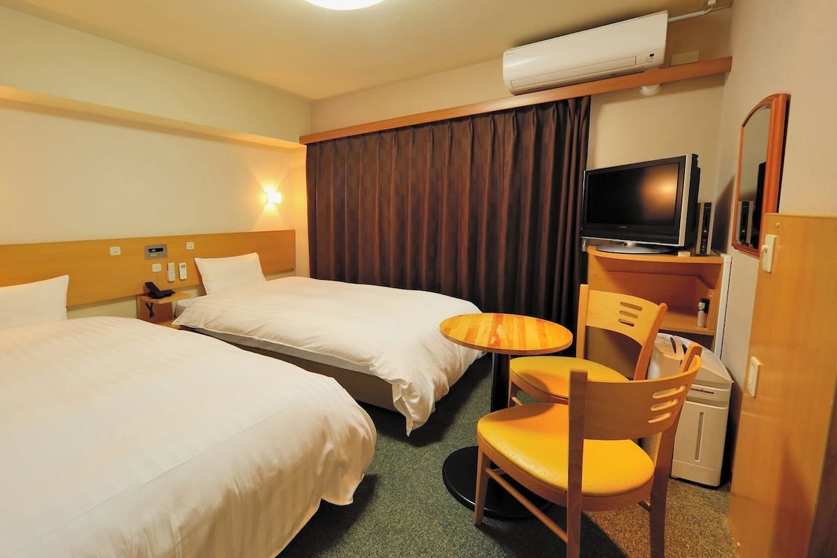 A hotel room with two beds, a desk with a chair, a television, and an air conditioning unit.