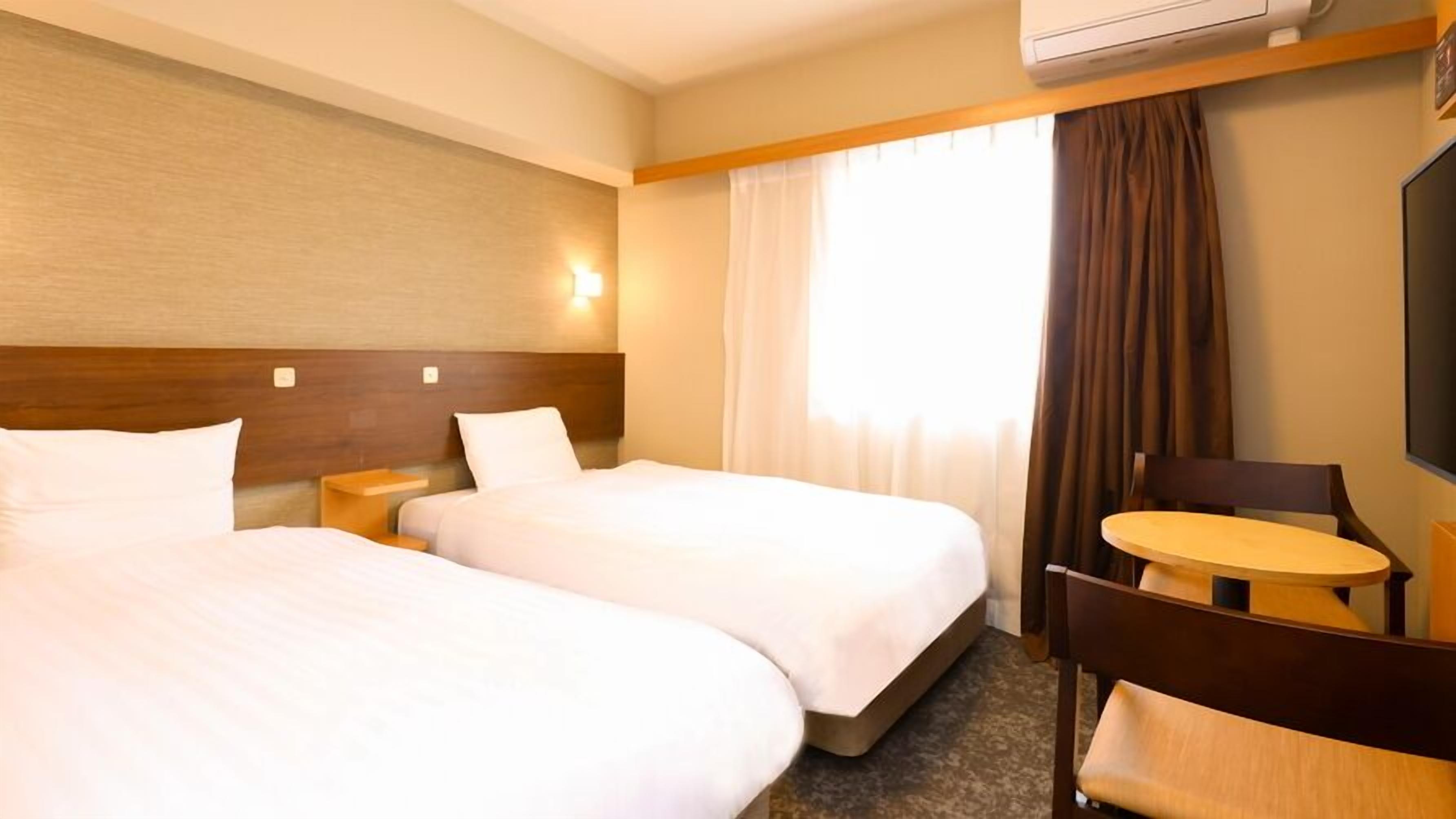 A hotel room with two beds, a small table, a chair, a TV, and an air conditioning unit.
