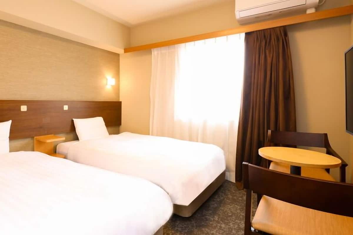 A hotel room with two beds, a small table, a chair, a TV, and an air conditioning unit.