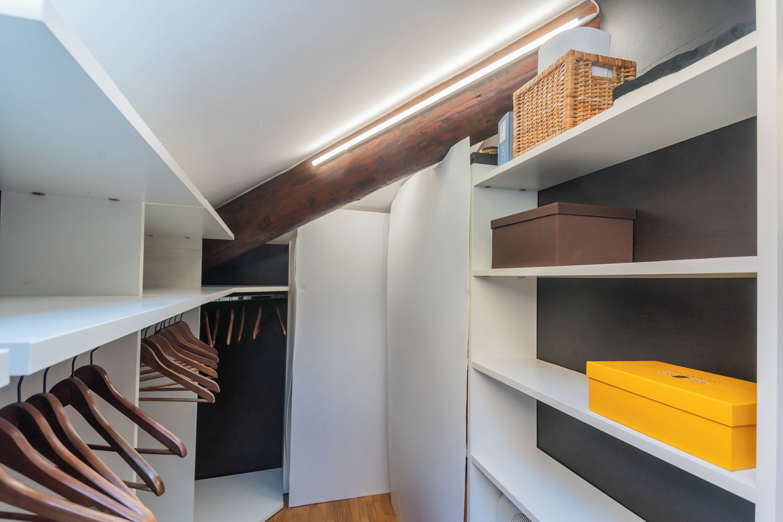 Un placard moderne avec des étagères blanches, une poutre en bois et une boîte jaune sur l’étagère supérieure.