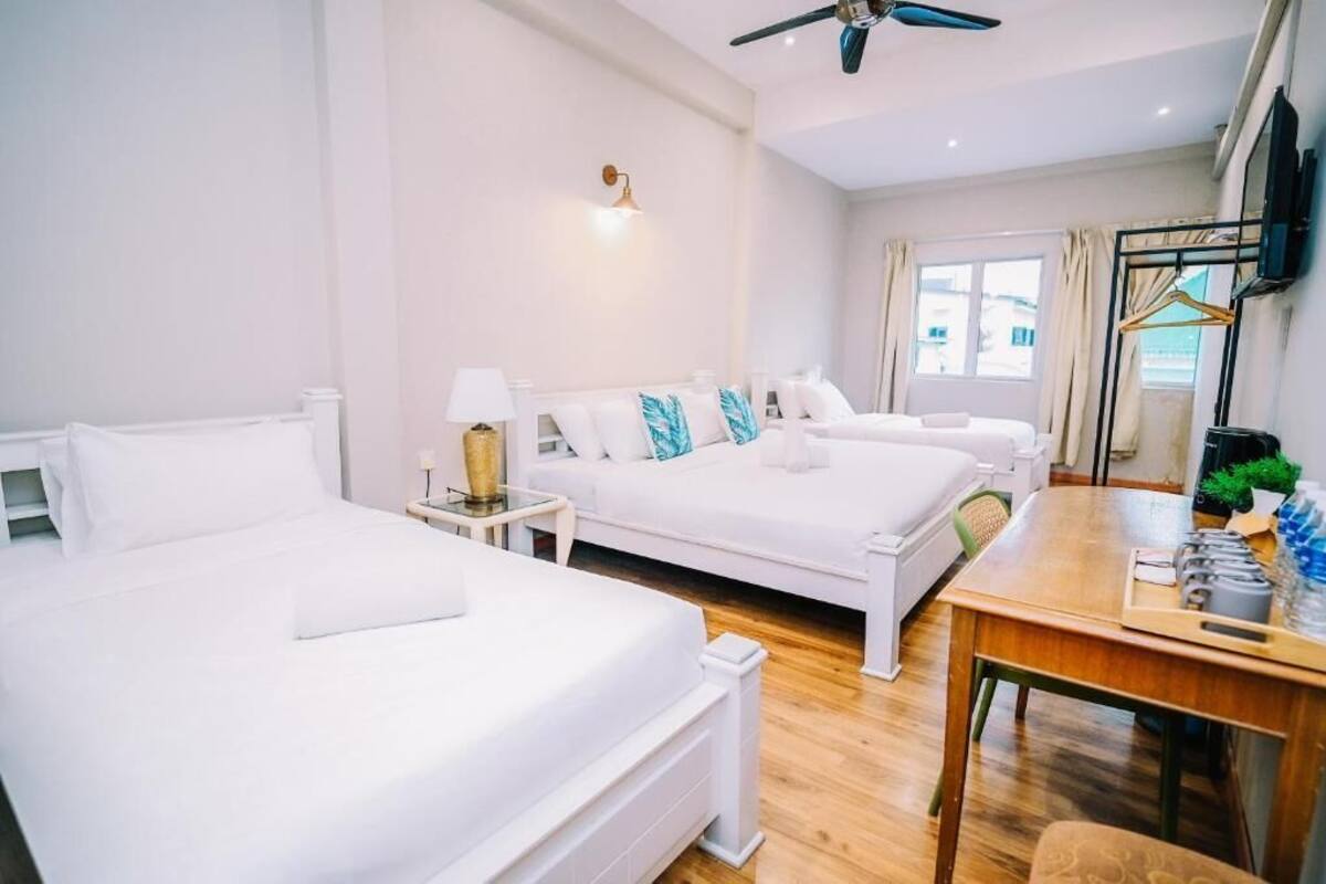 Habitación de hotel con dos camas, una mesa de madera, ventilador de techo, televisor y ventana con cortinas.