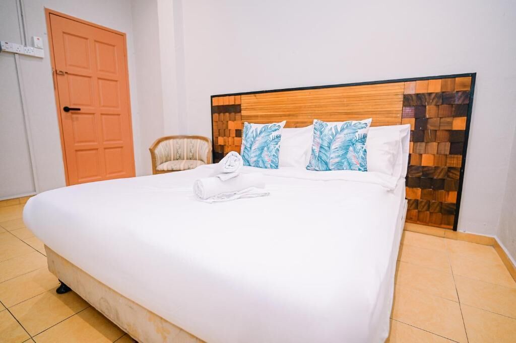 A neatly made bed with a wooden headboard, a rolled towel, and a chair in a simple room with a tiled floor and a wooden door.