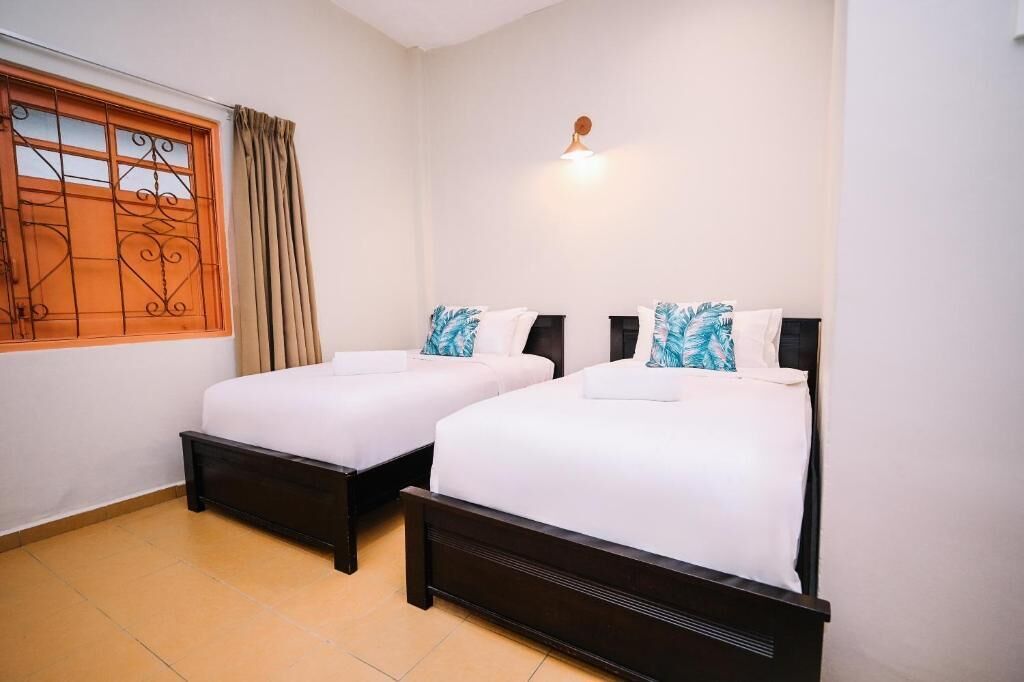 A hotel room with two beds, a window with curtains, a wall-mounted light, and a tiled floor.