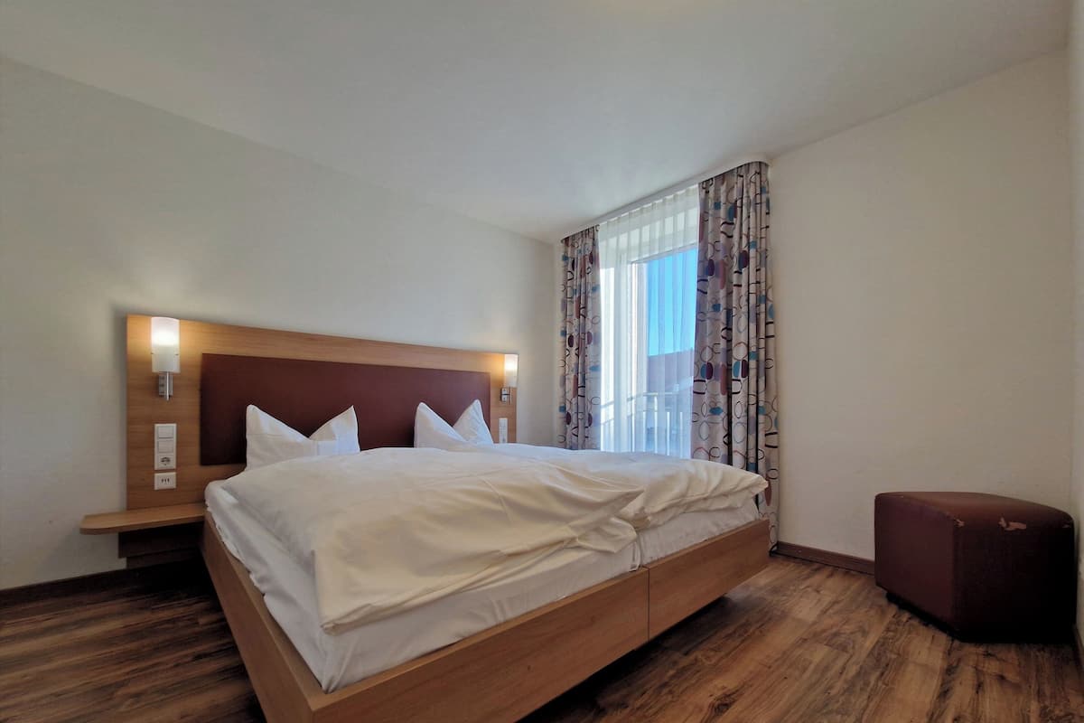 Une chambre d’hôtel avec un lit en bois, une table de chevet avec une lampe et une fenêtre ornée de rideaux à motifs.