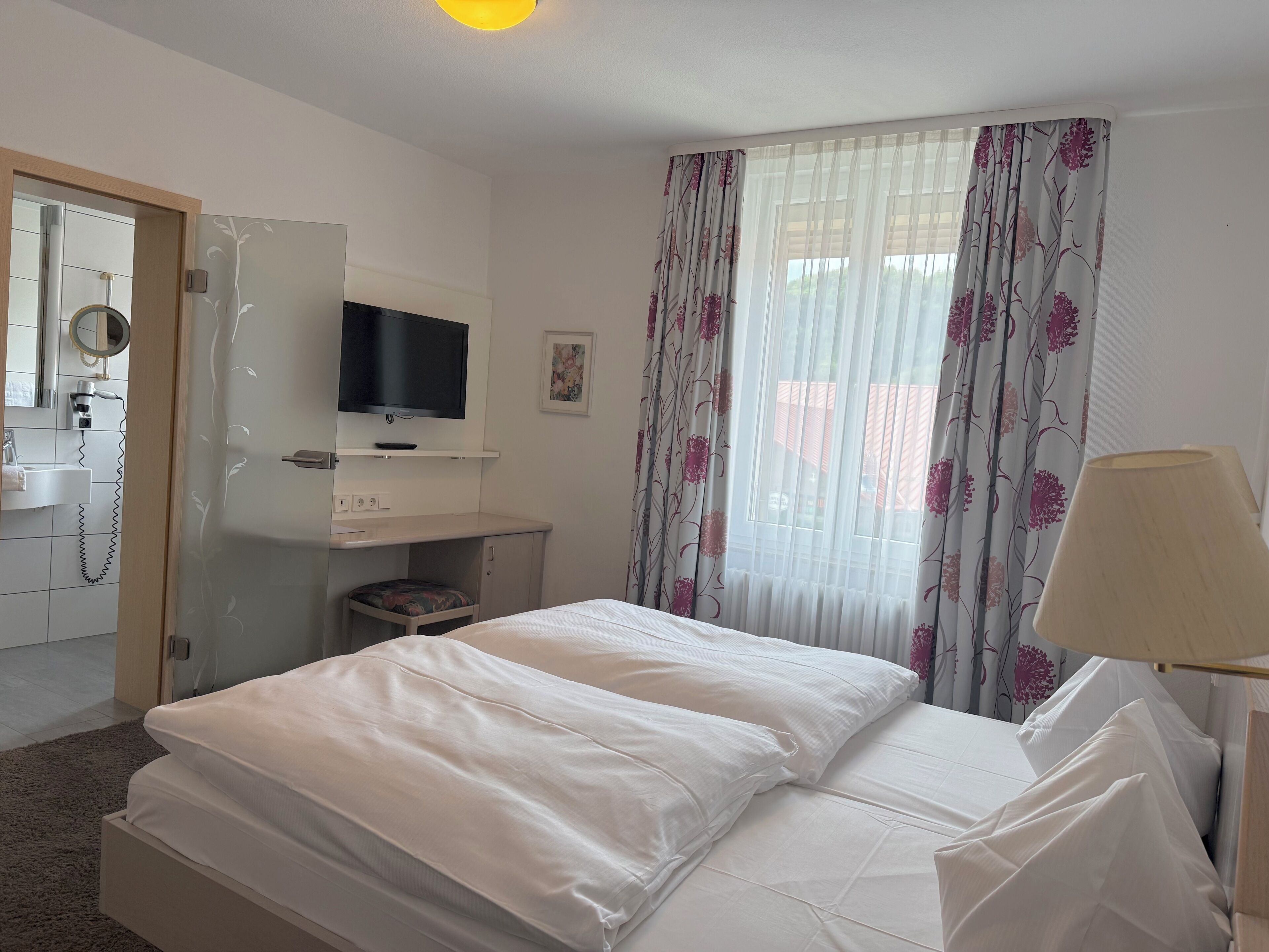 Quarto de hotel com uma cama, televisão na parede, secretária com cadeira, janela com cortinas florais e abajur na mesa de cabeceira.