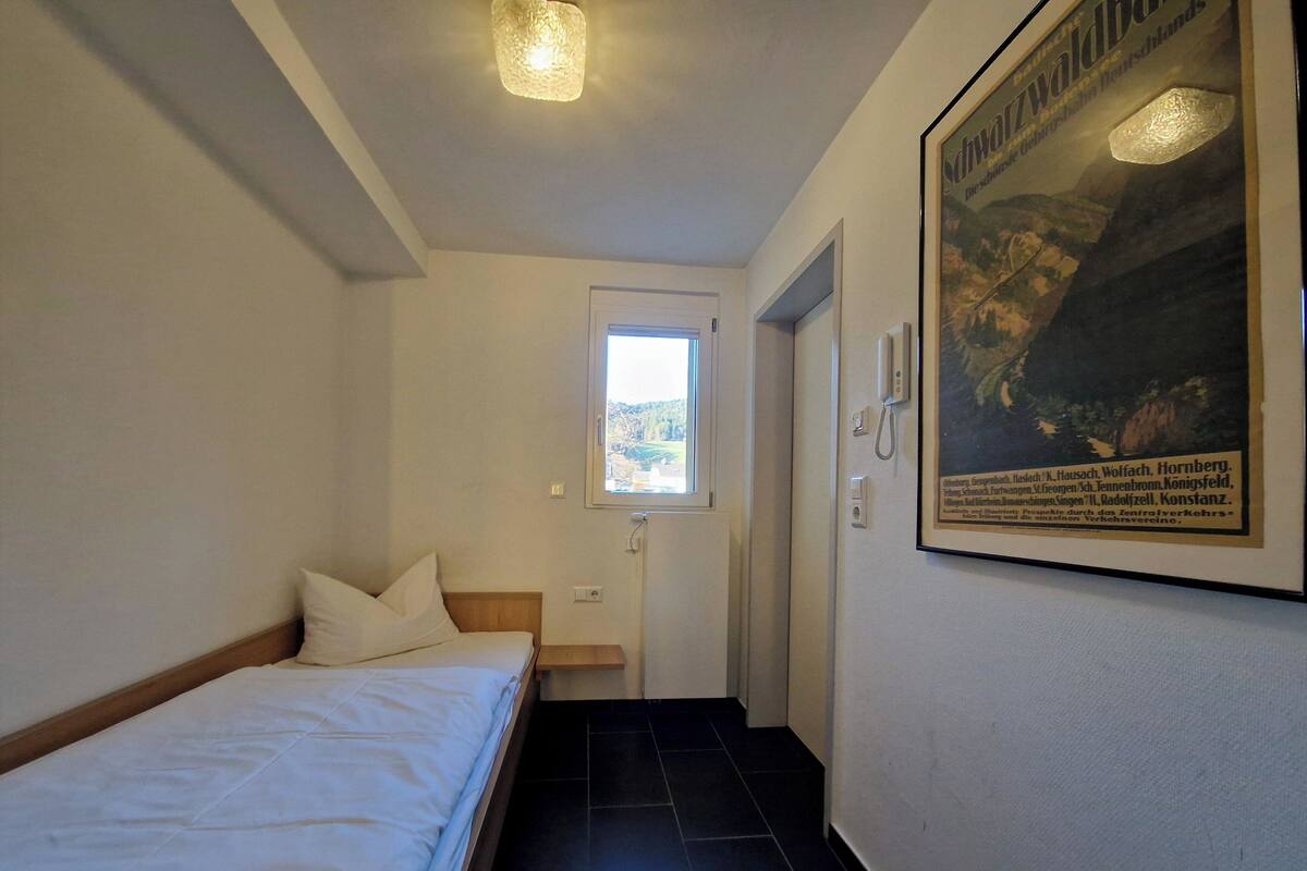 Une chambre d’hôtel avec un lit, une petite fenêtre, une porte et une affiche encadrée au mur.